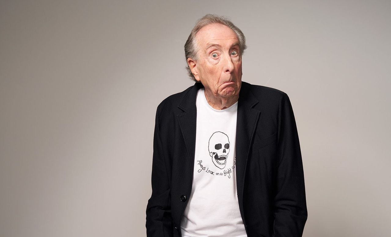 Comedian Eric Idle shrugs playfully while wearing a black blazer and a white graphic shirt reading Always Look on the Bright Side.